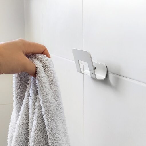 Mano colgando toalla en Gancho DOJO Mediano blanco Muett instalado en pared de baño, diseño funcional moderno minimalista