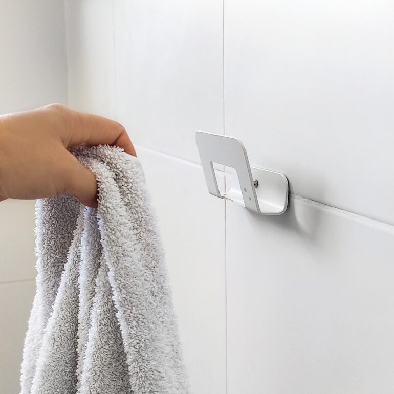 Mano colgando toalla en Gancho DOJO Mediano blanco Muett instalado en pared de baño, diseño funcional moderno minimalista